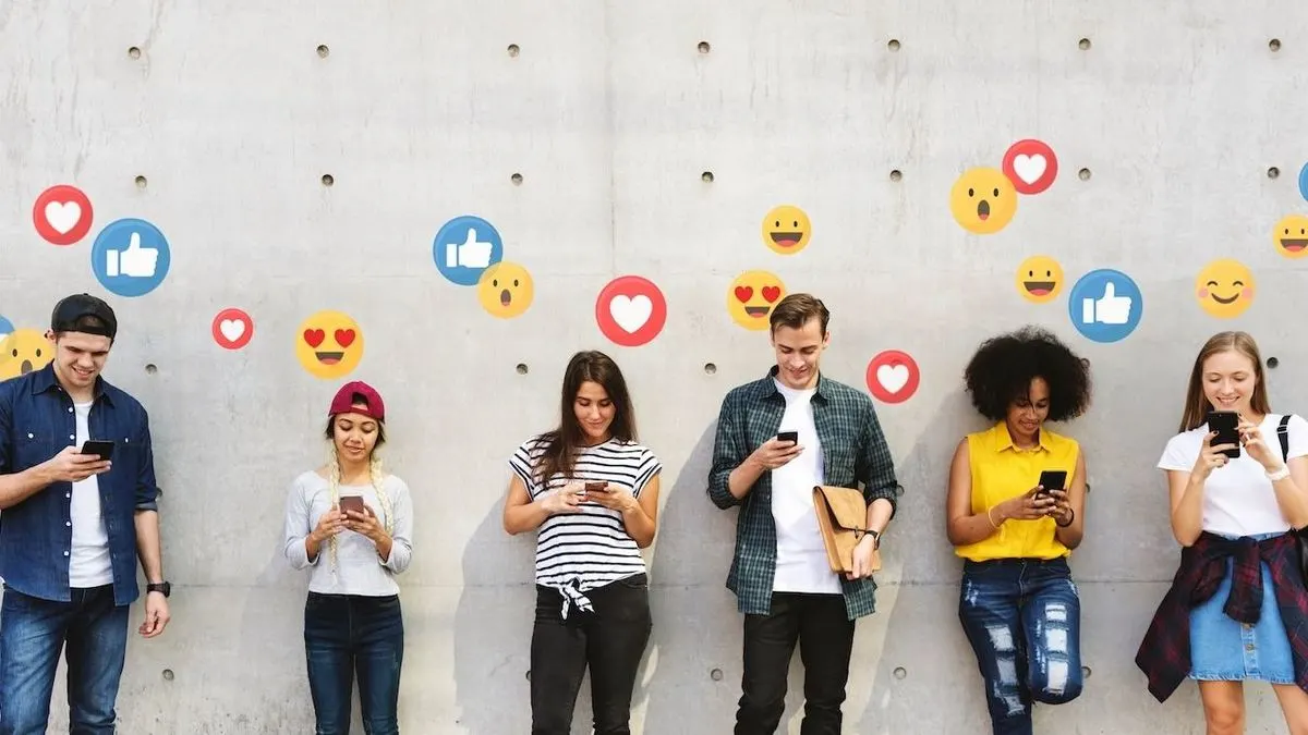 Sechs junge Leute stehen vor einer grauen Betonwand und blicken auf ihre Smartphones. Herzen, Daumen hoch und gelbe Emojis sind an der Wand verteilt.