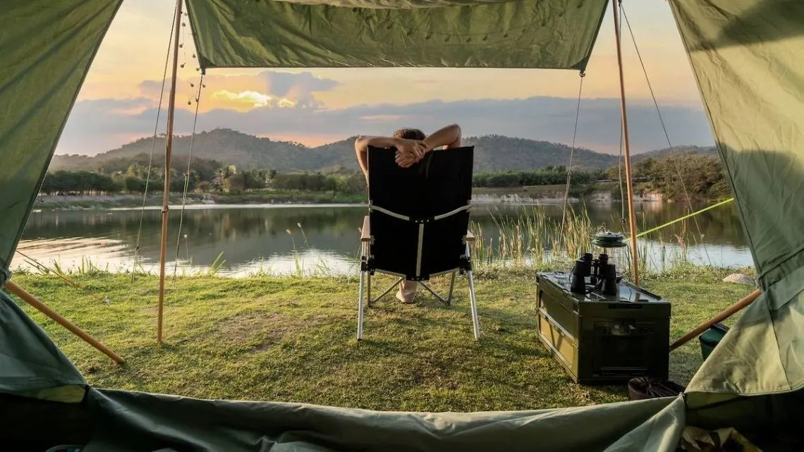 Blick aus einem grünen Zelt auf eine Person im Klappstuhl vor einem See mit Bergen bei Sonnenuntergang. Grüne Box mit Laterne und Fernglas.