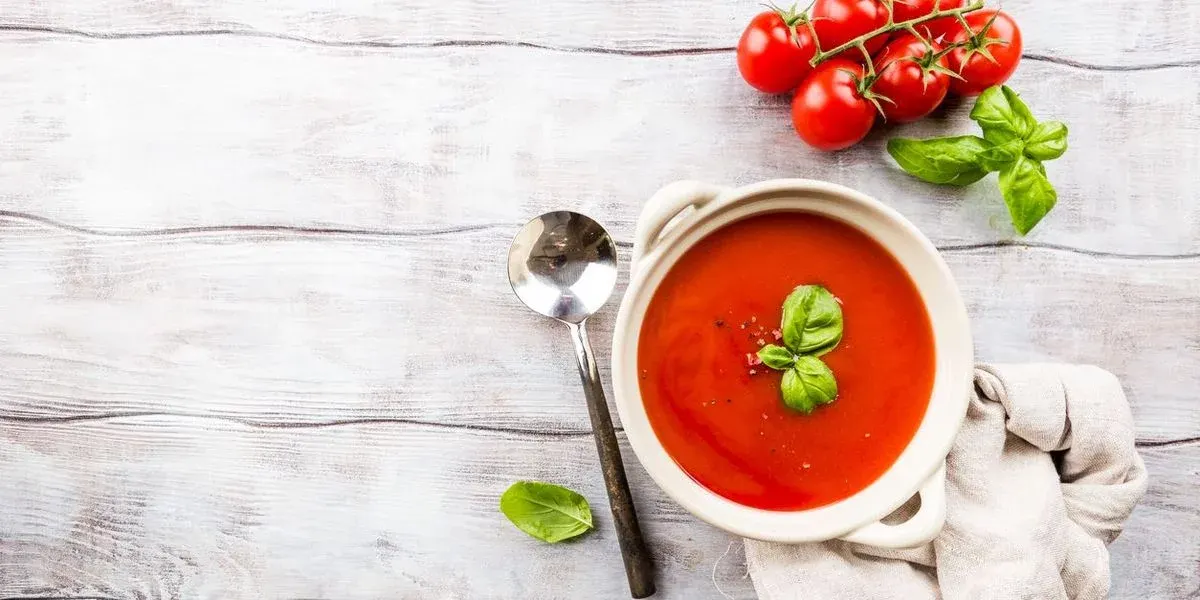 Eine Schale mit Gazpacho dekoriert mit Basilikum steht auf einem Holztisch neben einem Löffel und einer Tomatenrispe.