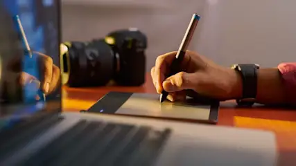 Hand hält silber-schwarzen Stift auf grauem Grafiktablett. Laptop-Tastatur vorne, schwarze Kamera hinten. Warme Beleuchtung.