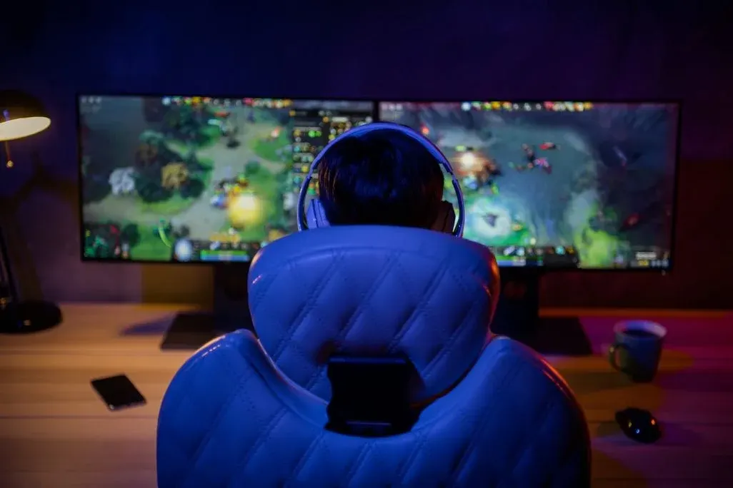 Gamer sitzt mit Headset in weißem Gaming-Stuhl vor zwei Monitoren und spielt ein buntes Online-Strategiespiel in einem abgedunkelten Zimmer.