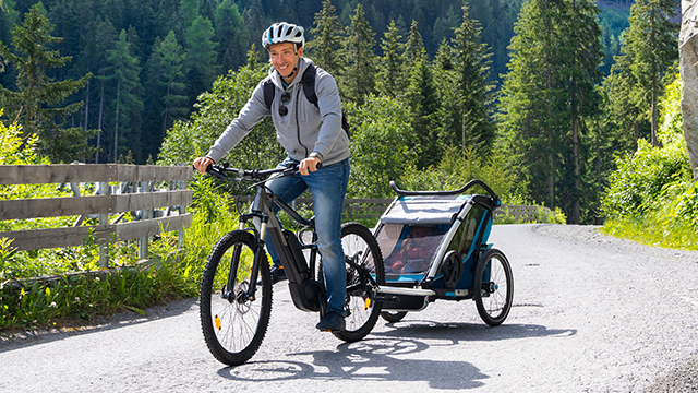 Ein Mann mit Helm fährt ein schwarzes Fahrrad mit blauem Anhänger auf einer Straße. Bäume im Hintergrund.