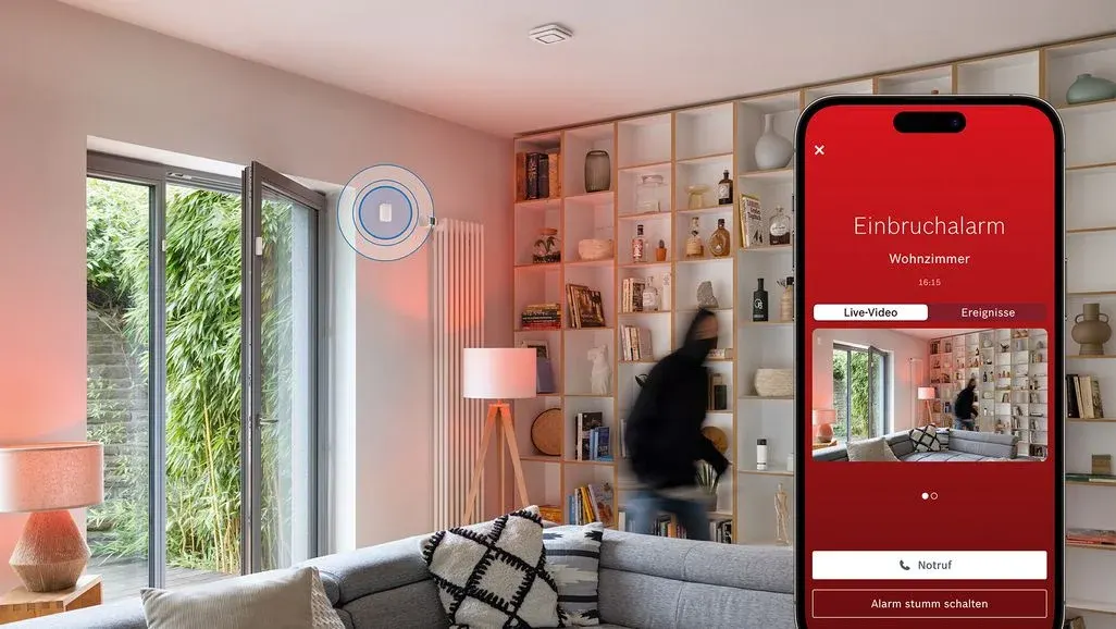 Modernes Wohnzimmer, offene Glastür zu Grün. Eine Person eilt durch rotes Licht. Sensor aktiv. Smartphone zeigt Einbruchalarm mit Live-Video des Raumes.