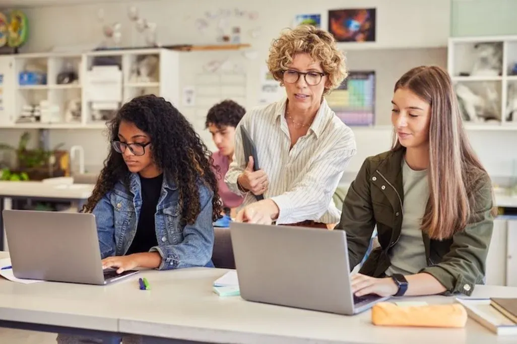 Lehrerin erklärt zwei Schülerinnen etwas am Laptop; beide arbeiten konzentriert im Klassenraum.
