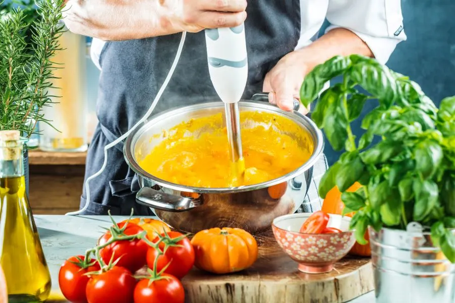 eine Person benutzt einen Stabmixer in einem Top der eine orange Masse beinhaltet, davor sieht man Tomaten, Öl und Basilikum
