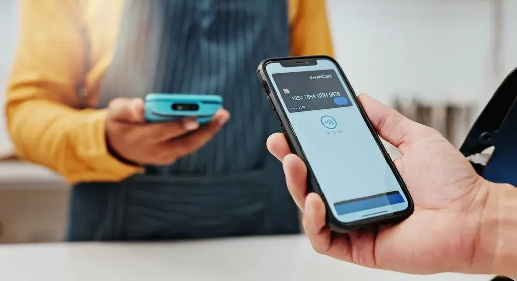 Kunde bezahlt kontaktlos mit Smartphone und digitaler Kreditkarte, Verkäufer hält mobiles Kartenterminal in der Hand.