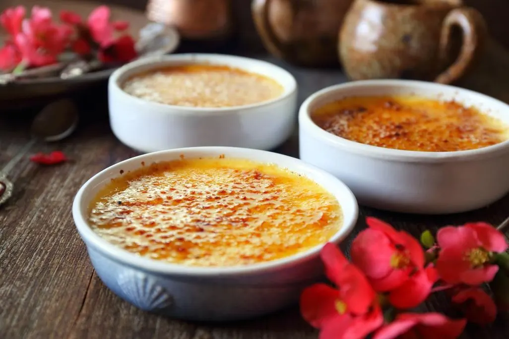 Drei Schalen mit Creme Crème Brûlée auf einem Holztisch, der mit roten Blüten dekoriert ist.