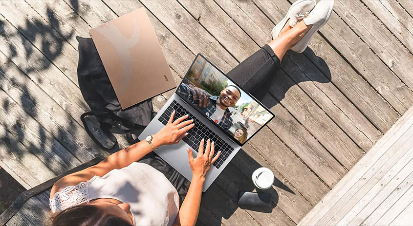 Eine Person sitzt auf einer Holzterrasse und tippt auf einem Laptop. Ein zweiter Laptop und eine Tasse stehen daneben.