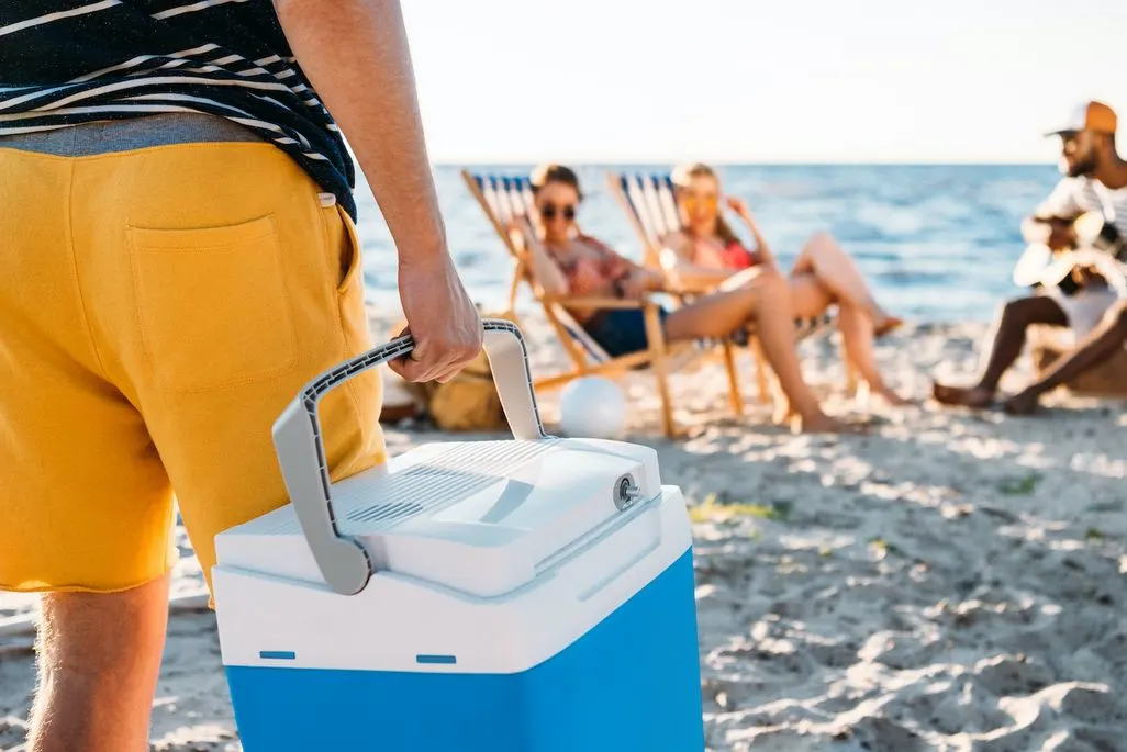Im Vordergrund eine Person die eine blaue Kühlbox am Strand trägt, im Hintergrund sitzen Leute.