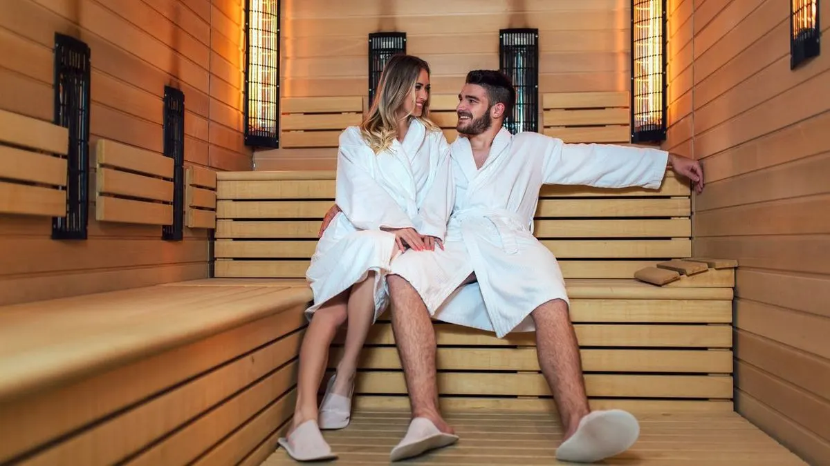 Ein Mann und eine Frau in weißen Bademänteln sitzen lächelnd auf Holzbänken in einer Sauna. Orange glühende Heizkörper sind an den Holzwänden.