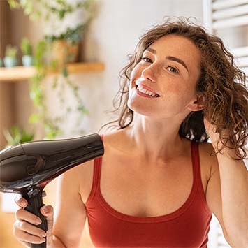 Frau mit Locken hält Föhn in der Hand. Rotes Top, Badezimmerhintergrund mit Pflanzen und Heizkörper.