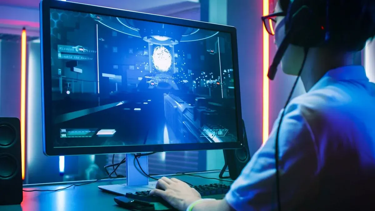 Spieler mit Kopfhörern spielt ein blaues Ego-Shooter-Videospiel an einem Monitor. Hände auf Tastatur und Maus. Orange/blaue Neonlichter im Raum.