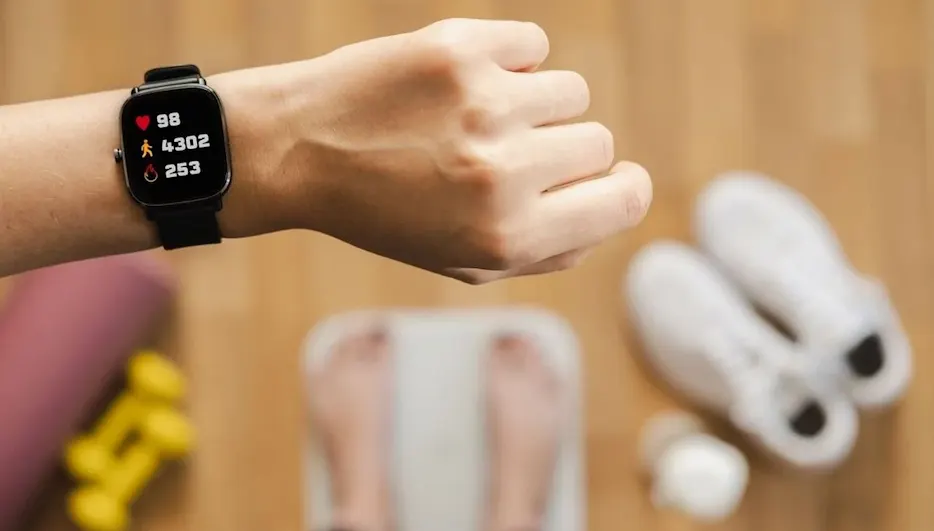 Person steht barfuß auf einer Waage, im Vordergrund zeigt eine Smartwatch am Handgelenk Puls, Schritte und Kalorien, daneben Sportschuhe und Hanteln.