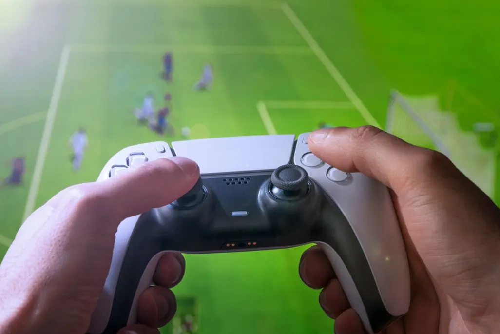 Hände halten einen PlayStation Controller, im Hintergrund läuft ein Fußballspiel auf dem Bildschirm.