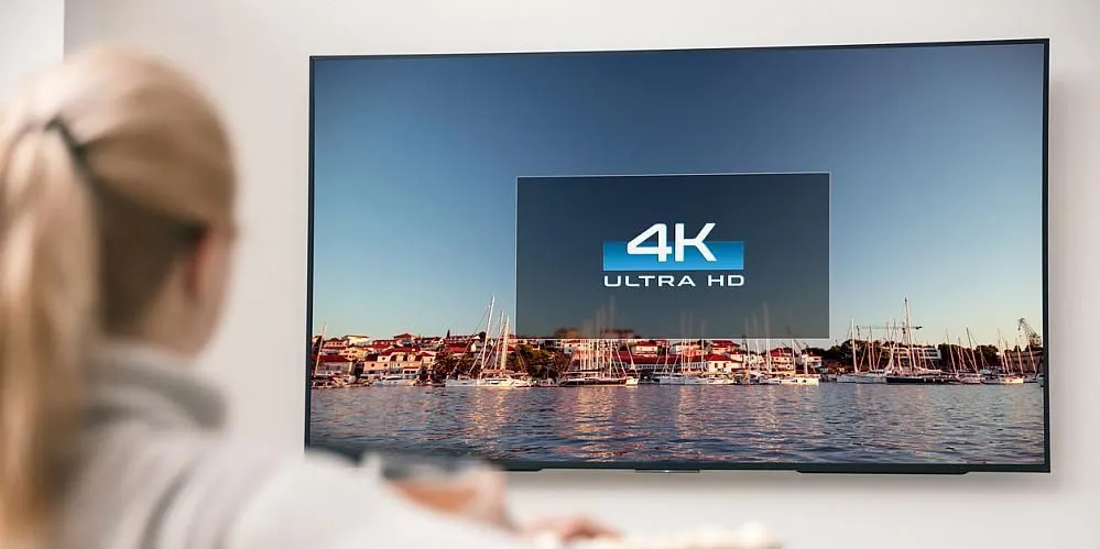 Frau sitzt vor einem Fernseher mit 4K Ultra HD Logo und Blick auf einen Hafen mit Segelbooten.