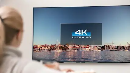Frau sitzt vor einem Fernseher mit 4K Ultra HD Logo und Blick auf einen Hafen mit Segelbooten.