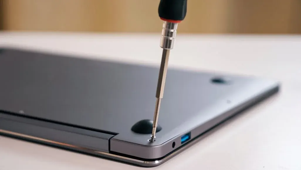 Ein silberner Schraubendreher mit schwarzem Griff löst eine Schraube am grauen Laptop. Ein schwarzer Gummifuß und blauer USB-Anschluss sind sichtbar.