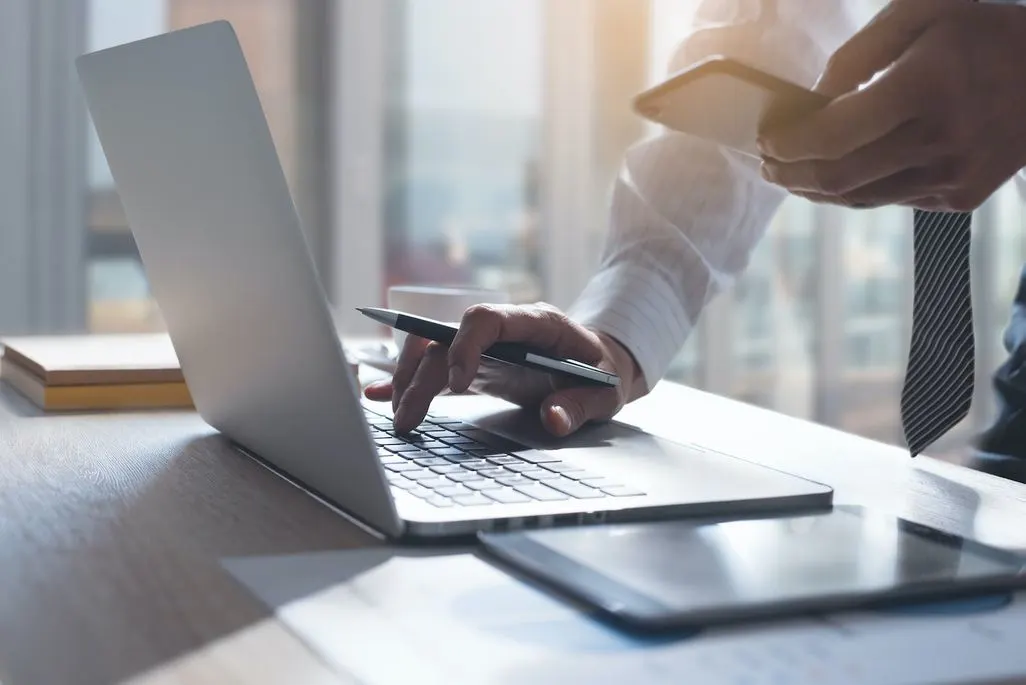 Eine Person tippt am Laptop während sie ein Smartphone in der anderen Hand hält.