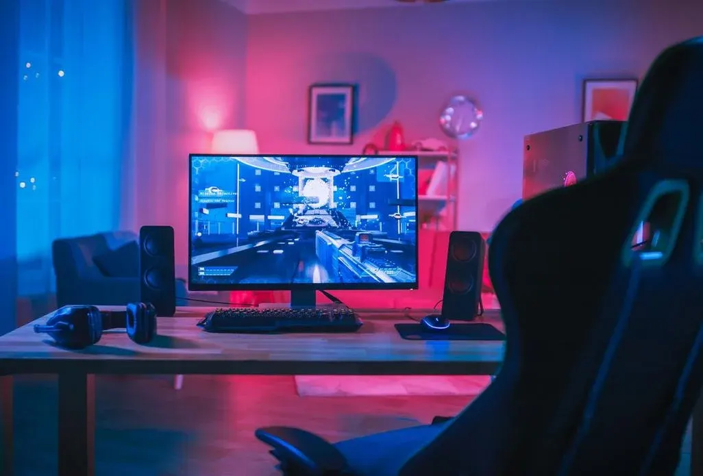 In einem rosa-blau beleuchteten Gaming-Zimmer steht ein Monitor auf einem Gaming-Tisch.  