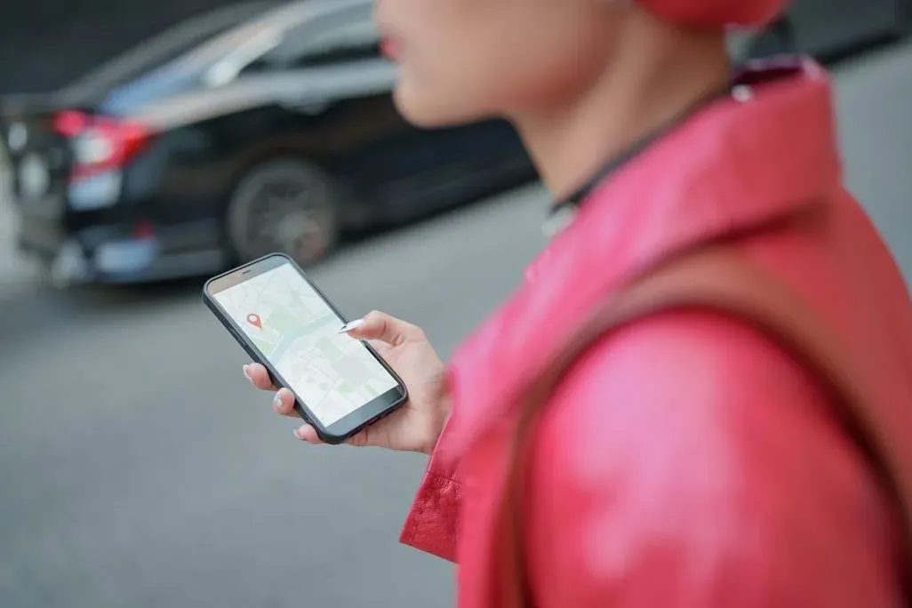 Person in pinker Jacke hält schwarzes Smartphone mit Karten-App und rotem Pin, tippt. Unscharfes schwarzes Auto im Hintergrund.