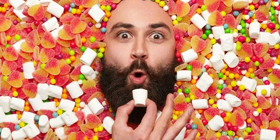 Ein bärtiges Gesicht ragt aus einem bunten Berg von Herzgummis, kleinen Bonbons und Marshmallows. Eine Hand hält ein Marshmallow zum Mund.