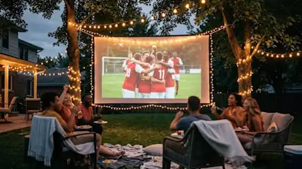 Freunde sehen ein Fußballspiel auf einer Leinwand im grünen Garten mit Lichterketten. Sie sitzen, essen und jubeln.