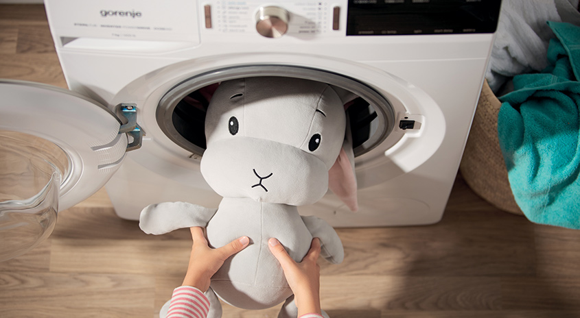 Ein Kind legt ein graues Stofftier-Kaninchen in eine weiße Waschmaschine. Ein Wäschekorb steht daneben.