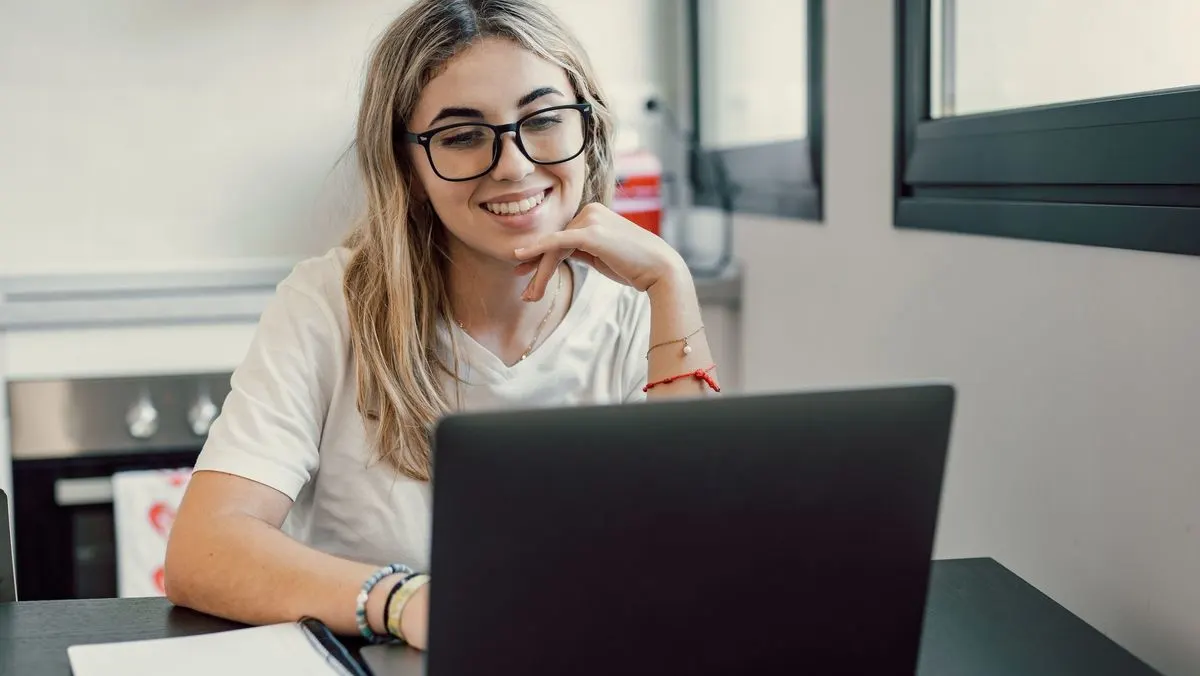 Eine lächelnde Person mit Brille und weißem T-Shirt hält die Hand am Kinn, blickt auf den Laptop. Notizbuch auf Tisch. Küche und Fenster im Hintergrund.