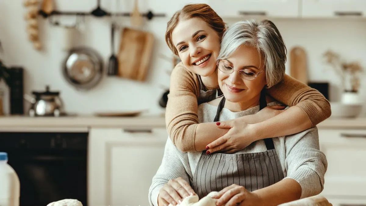Jüngere Person umarmt lächelnd ältere Person, die in gestreifter Schürze Teig knetet. Eine Küche mit Kochutensilien im Hintergrund.