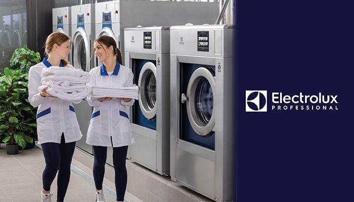 2 Frauen in einer Wäscherei mit Handtüchern in der Hand, Waschmaschinen und Trockner im Hintergrund. Rechts befindet sich das Electrolux Logo