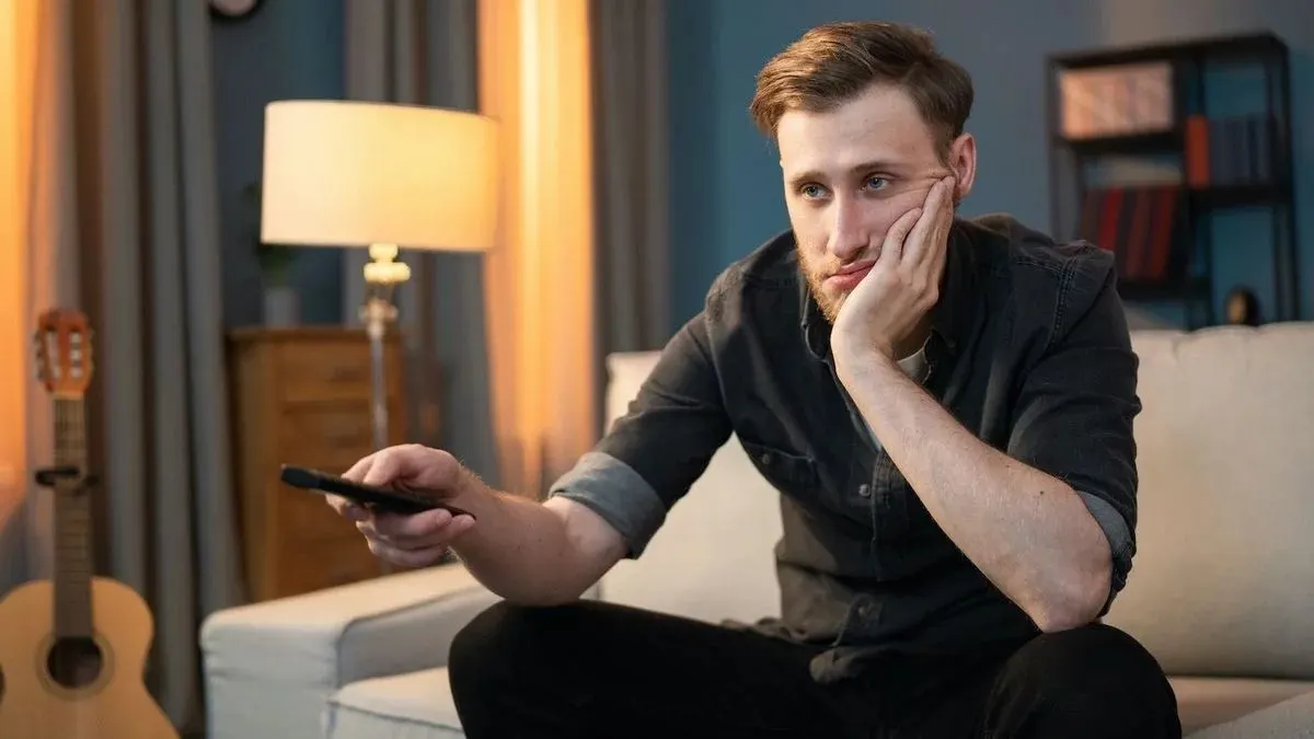 Ein Mann mit genervtem Gesichtsausdruck sitzt auf einem Sofa und hält eine Fernbedienung.