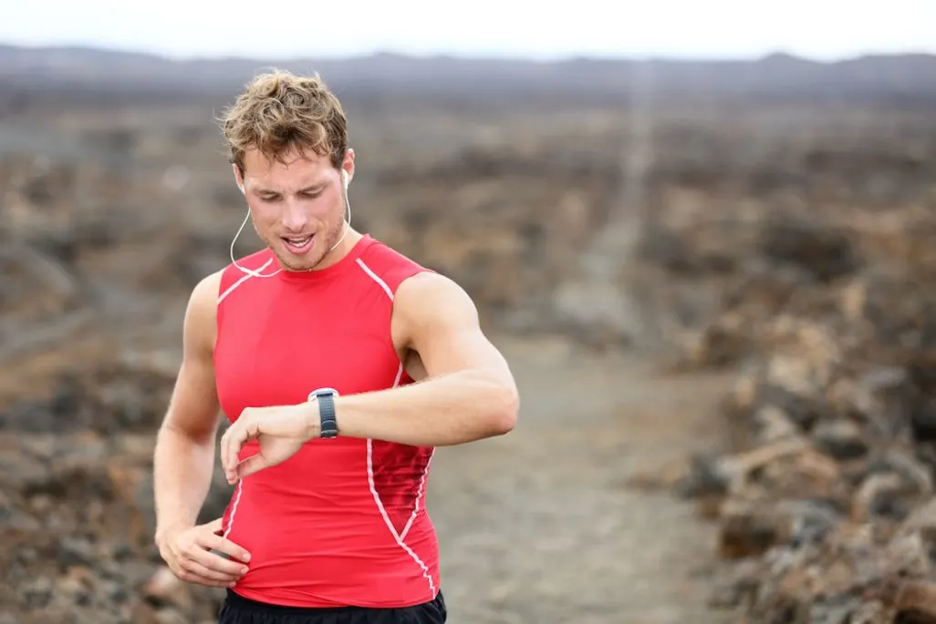 Ein Mann in rotem Sportshirt trägt eine Smartwatch und steht vor einer bergigen Landschaft.