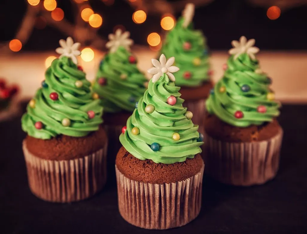 Cupcakes mit grünem Tannenbaum-Frosting und bunten Zuckerperlen vor Lichterdekoration.