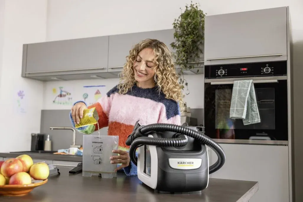 Lächelnde Frau in buntem Pullover füllt Flüssigkeit in einen Kärcher Reinigungsbehälter. Moderne Küche mit Backofen, Schränken und Äpfeln.