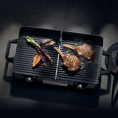 Auf einem schwarzen Grill liegen zwei Steaks mit Knochen und Gemüse. Spargel, Zwiebeln und eine Scheibe Kürbis liegen auf der linken Seite.