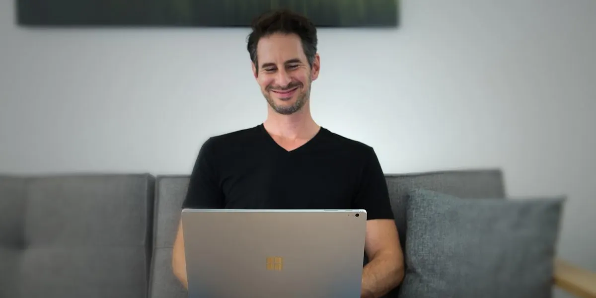 Ein lächelnder Mann in schwarzem Shirt sitzt auf einem grauen Sofa und blickt auf einen silbernen Laptop. Im Hintergrund ist eine helle Wand mit einem dunklen Bild.
