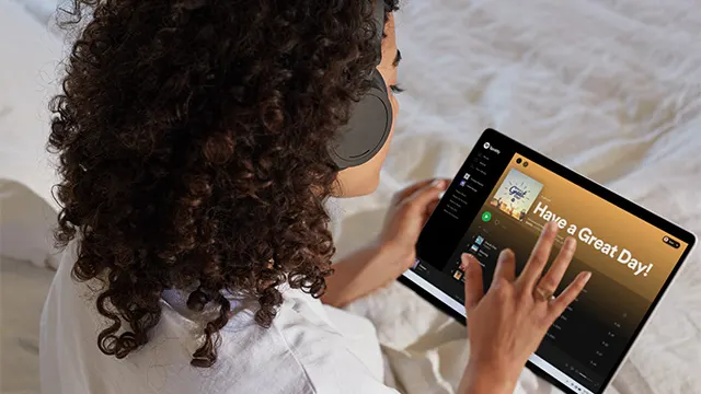 Frau mit Kopfhörern im Bett, die ein Tablet mit der Aufschrift "Have a Great Day!" benutzt.