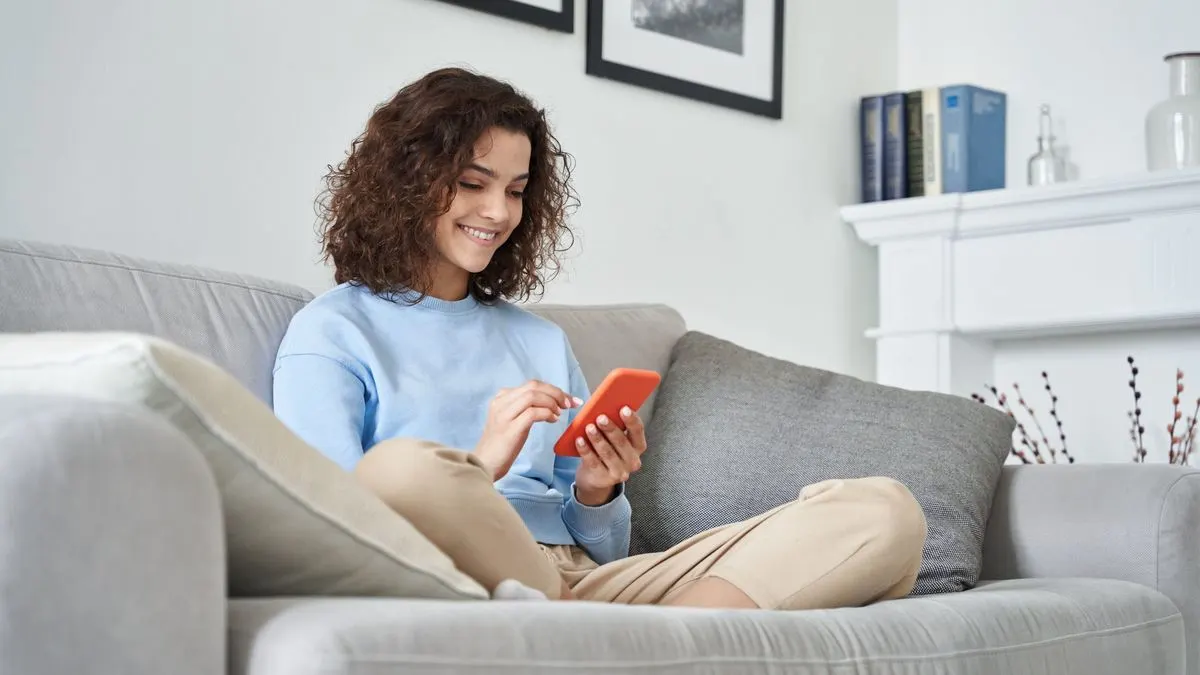 Person auf hellgrauem Sofa, in hellblauem Oberteil, tippt auf oranges Smartphone. Hintergrund: Bilder, Kaminsims mit Büchern und Vasen.
