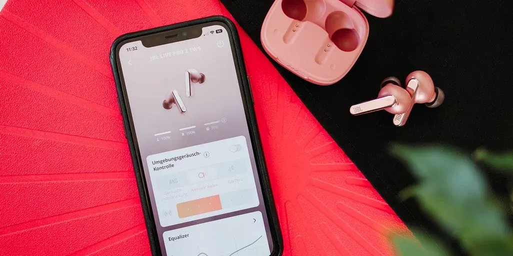 Ein Smartphone zeigt die Steuerung der rosegoldenen JBL Live Pro 2 TWS Ohrhörer-App. Dazu liegen die Ohrhörer und ihr Ladecase.