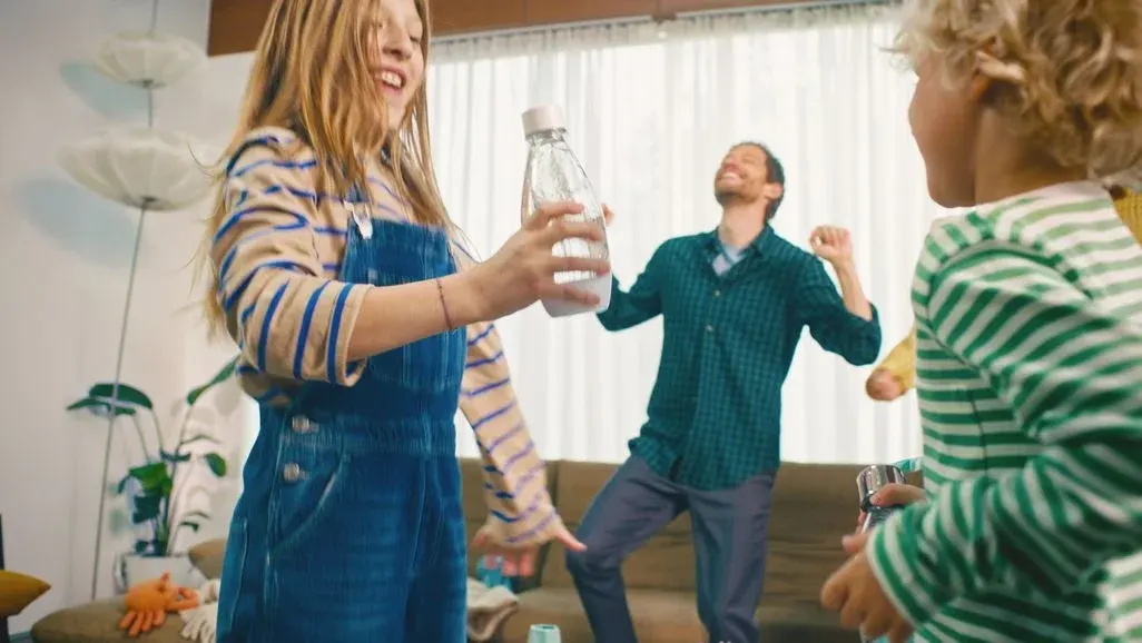 Ein Mädchen in Latzhose hält eine Flasche, während ein Mann im karierten Hemd und ein Junge im gestreiften Shirt fröhlich in einem Wohnzimmer tanzen.