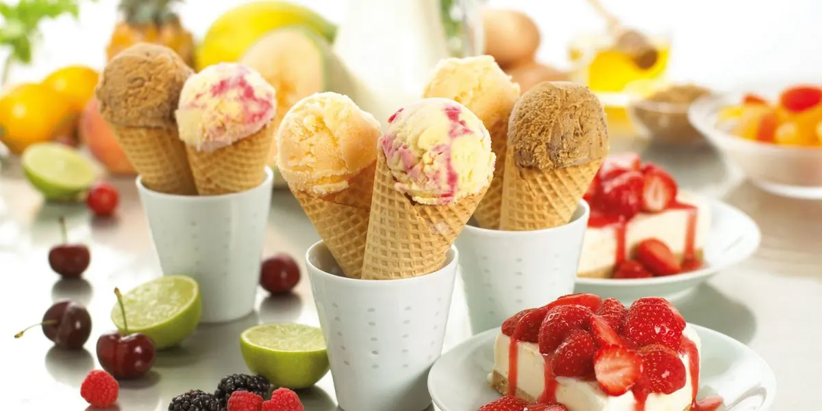 Vier Waffeleiswaffeln mit verschiedenen Eissorten in Bechern und Erdbeerkuchen im Hintergrund.