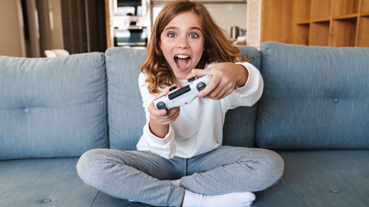 Auf einem blaugrauen Sofa sitzt ein Mädchen und spielt Videospiele mit einem weißen Controller und blickt überrascht. 