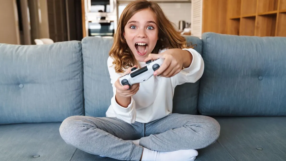 Auf einem blaugrauen Sofa sitzt ein Mädchen und spielt Videospiele mit einem weißen Controller und blickt überrascht.
