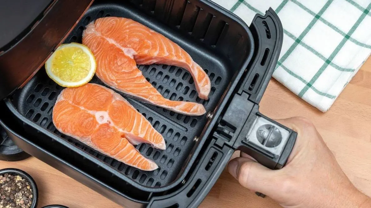 Zwei rohe Lachsfilets und Zitrone in schwarzem Frittierkorb, eine Hand zieht ihn. Weißes Tuch mit grünen Karos, Holzfläche.