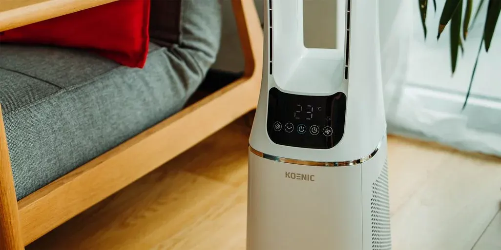 Weißer KOENIC Luftreiniger mit "23°C" Anzeige, neben Holz-Sessel mit grauem Polster und rotem Kissen, am Fenster mit Pflanze.
