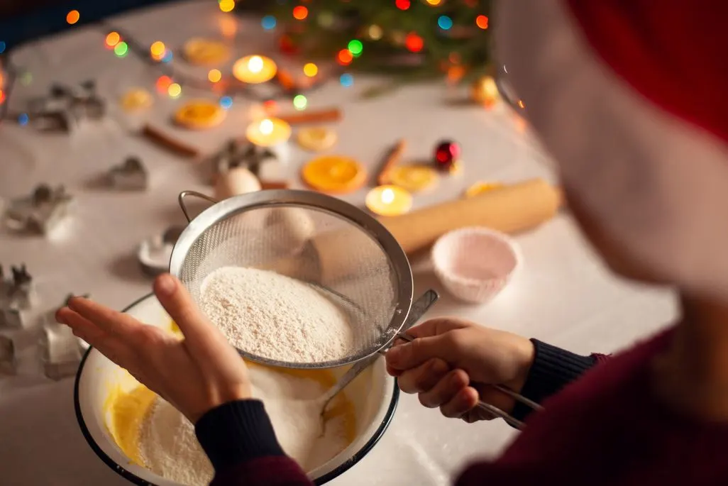Person siebt Mehl in eine Schüssel beim Weihnachtsbacken mit Deko im Hintergrund.