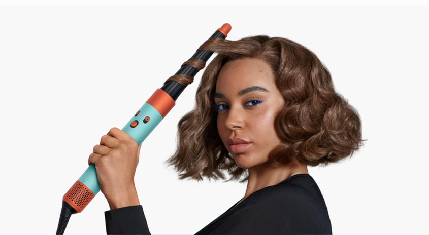 Eine türkisfarbene und orangefarbene Haarsträhne wird zum Locken von braunem Haar verwendet, wobei ein schwarzes Kabel sichtbar ist.