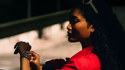 Person mit roter Jacke, weiße Sonnenbrille auf dem Kopf. Blickt auf beleuchtete Apple Watch 11 am linken Handgelenk vor dunklem Hintergrund.