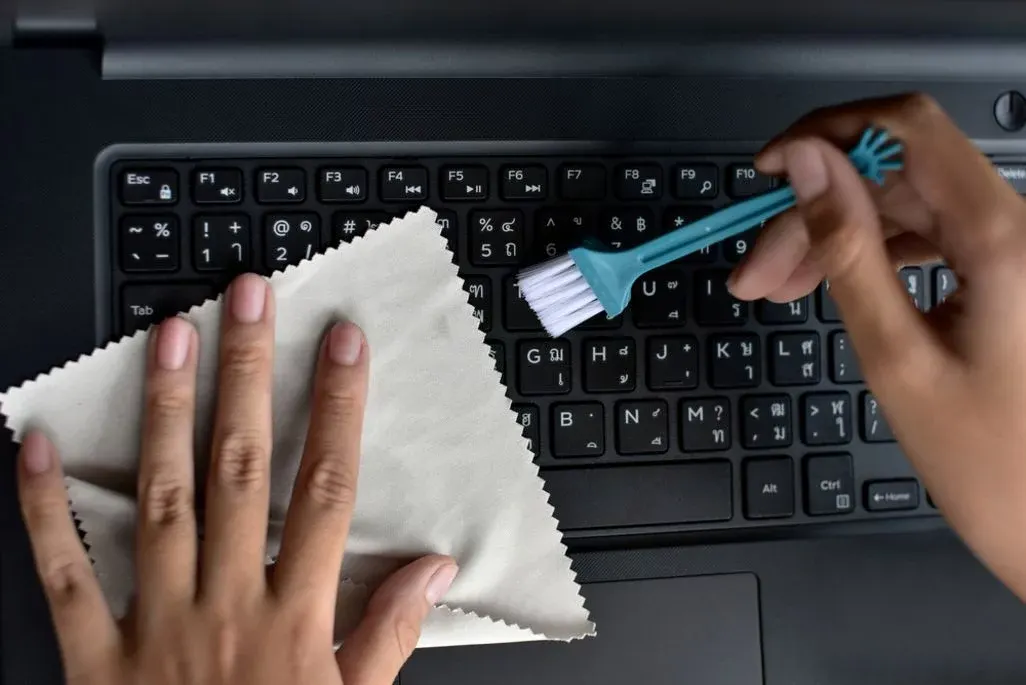 Hände reinigen eine schwarze Laptop-Tastatur mit einem beigen Tuch und einer blauen Bürste.