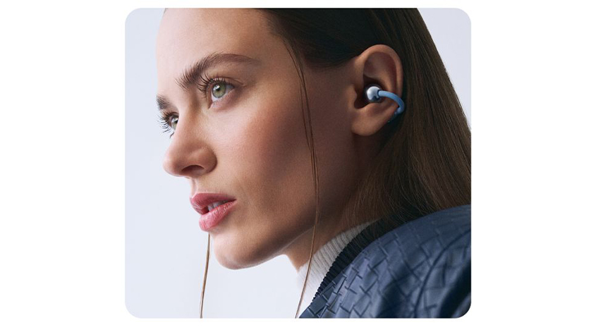  Nahaufnahme einer Person im Profil mit im Ohr sitzenden, blauen In‑Ear-Kopfhörern, vor hellem Hintergrund.